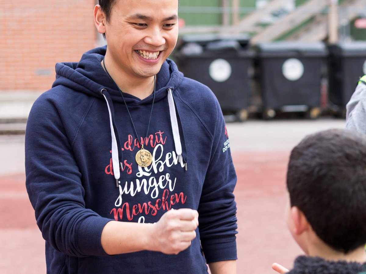 Lächelnder Mann in blauem Hoodie mit Medaillon interagiert mit Kind in grünem Hoodie auf Spielplatz.