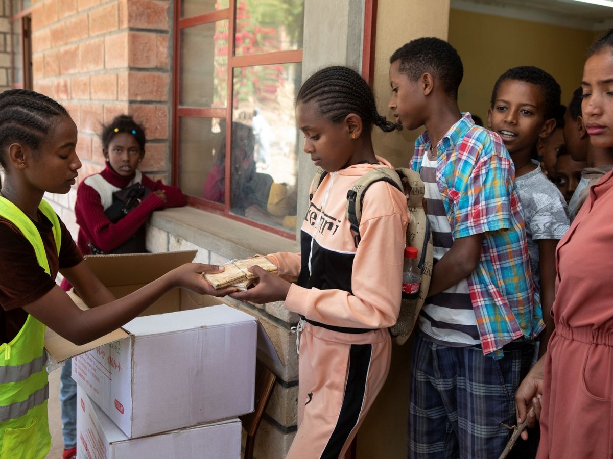 Eine junge Freiwillige übergibt einem Mädchen Holz-Lernmaterialien aus einer Reihe wartender Schüler vor einem Schulgebäude.