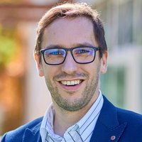 Nahaufnahme eines lächelnden Mannes mit Brille und hellbraunem Haar, der eine dunkle Jacke trägt.