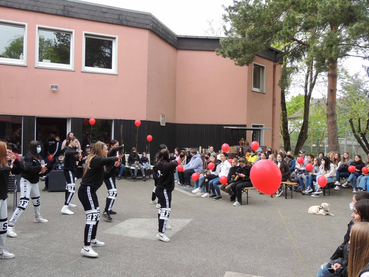 Outdoor-Tanzaufführung einer Jugendgruppe in Trainingsanzügen, beobachtet von Zuschauern mit roten Ballons vor einem rosa Gebäude.