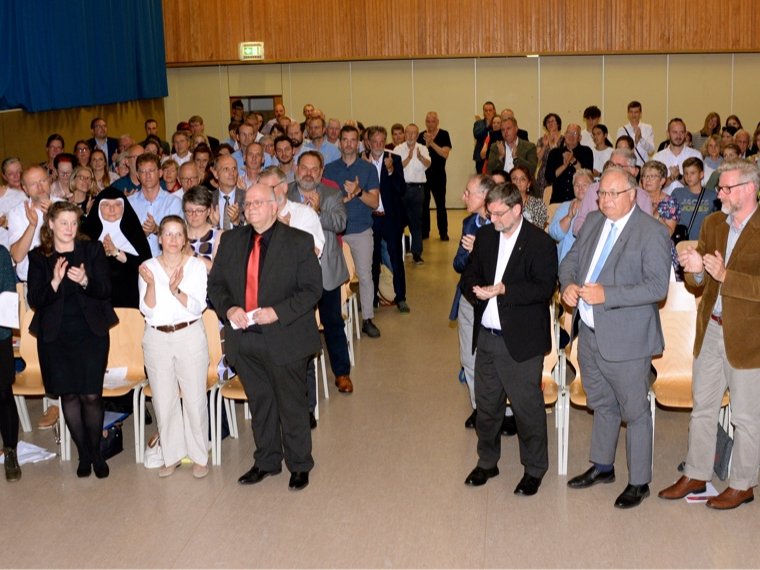 Zahlreiche Personen, darunter Männer und Frauen, stehen in einem großen Saal und applaudieren, teilweise in den ersten Reihen.