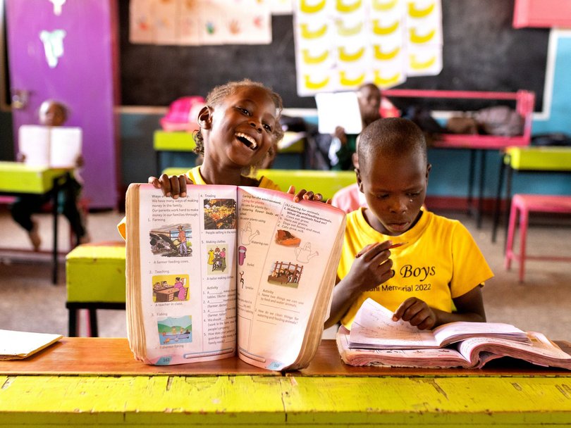 Ein fröhliches afrikanisches Mädchen hält ein offenes Lehrbuch und lacht, während ein Junge in einem lebhaften Klassenzimmer lernt.