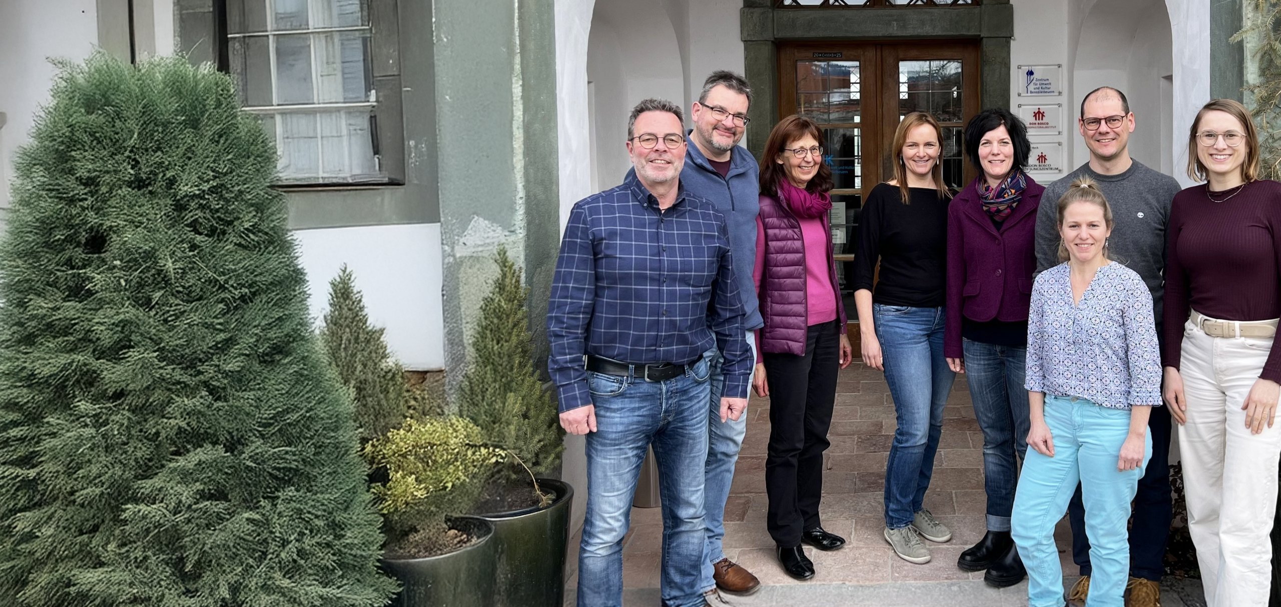Eine lächelnde Gruppe von neun Personen, fünf Frauen und vier Männern, steht vor einem Gebäude mit Torbögen und grünen Pflanzen.