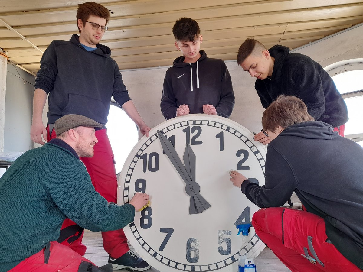 Fünf Personen, darunter junge Männer und ein älterer Mann, reinigen ein großes Zifferblatt mit schwarzen Zahlen und dunkelgrauen Zeigern.