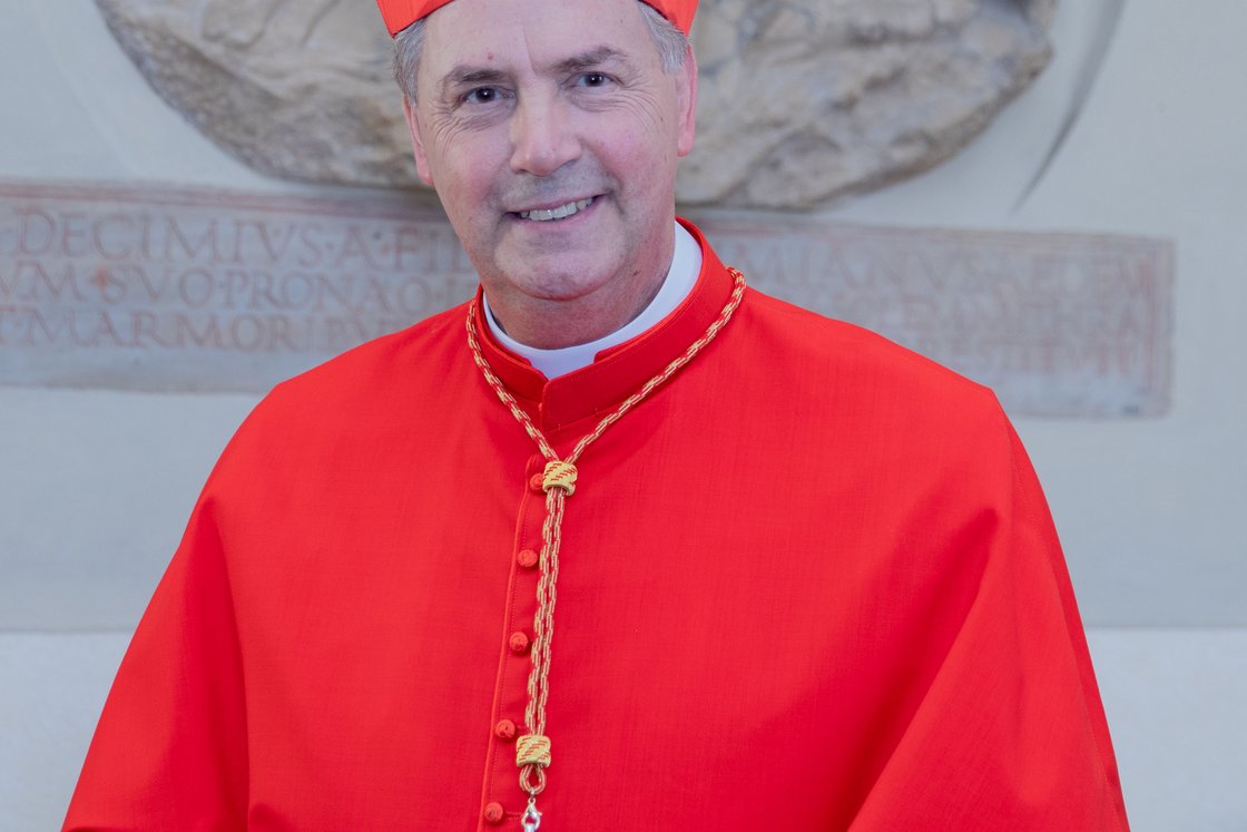 Lächelnder Kardinal in roter kirchlicher Kleidung und Birett, ein Pektoralenkreuz tragend, vor einem Steinrelief.