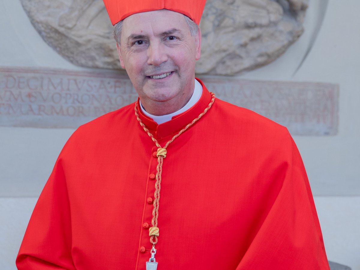 Lächelnder Kardinal in roter kirchlicher Kleidung und Birett, ein Pektoralenkreuz tragend, vor einem Steinrelief.