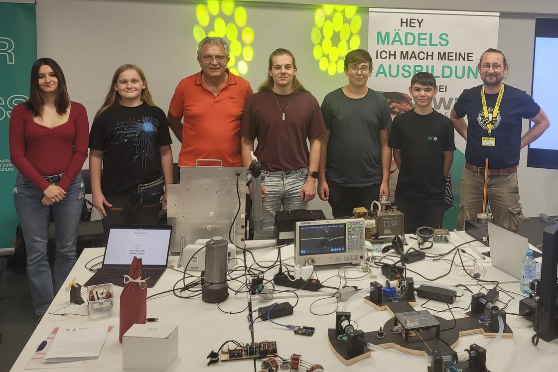 Zwei Frauen und fünf Männer/Jungen präsentieren am Messestand Technologieprojekte mit Elektronik, Experimenten und Laptops.