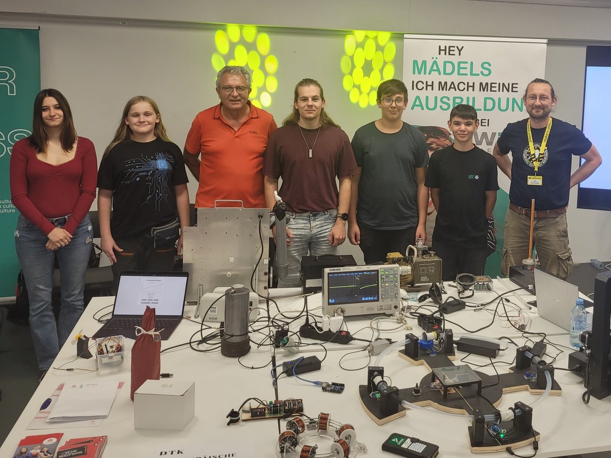 Zwei Frauen und fünf Männer/Jungen präsentieren am Messestand Technologieprojekte mit Elektronik, Experimenten und Laptops.