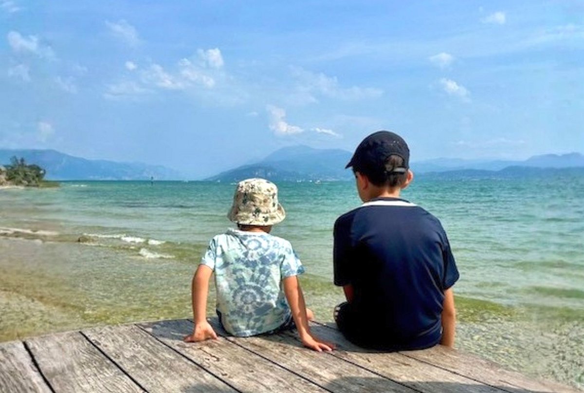 zwei Kinder sitzen auf Steg und schauen auf See