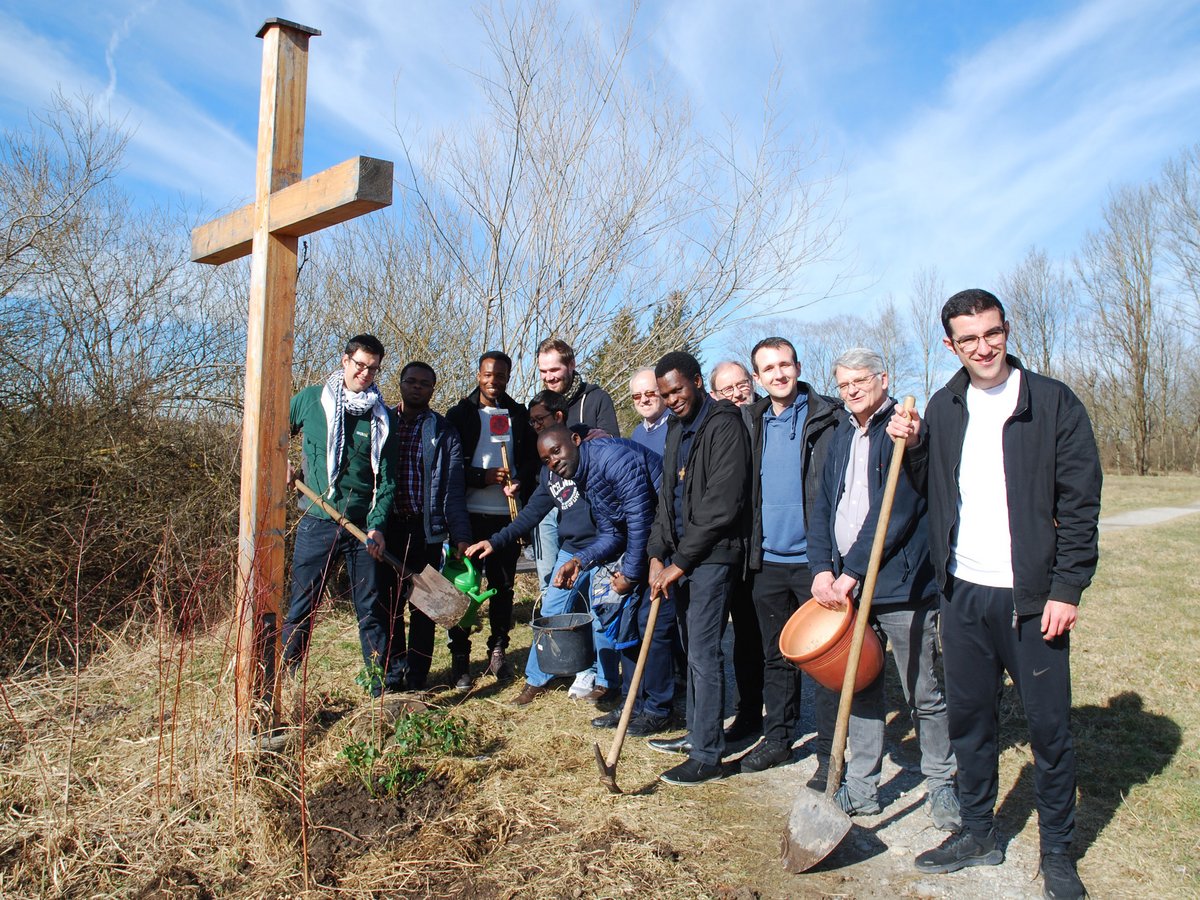 Vielfältige Gruppe von Männern pflanzt eine Pflanze am Fuße eines großen Holzkreuzes, hält Gartenwerkzeuge und lächelt.