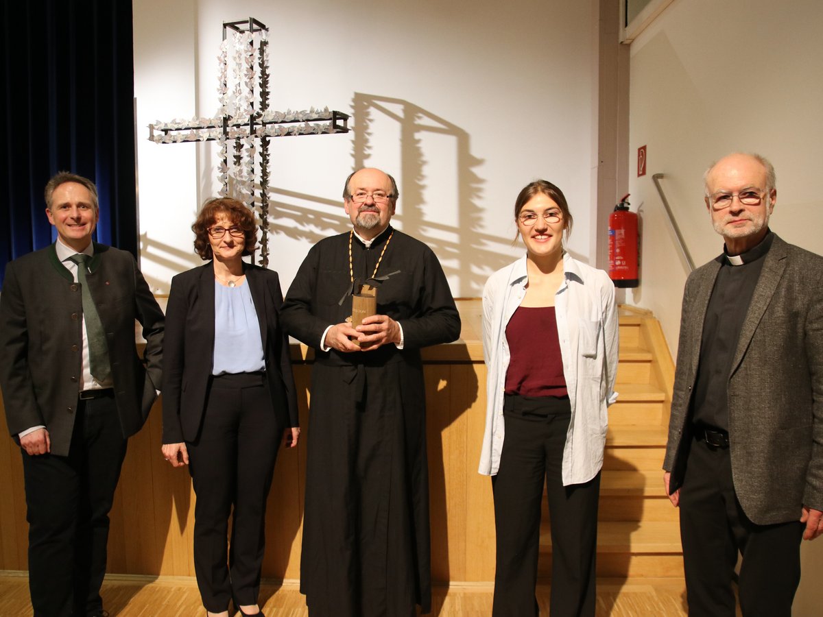 Fünf Personen, darunter zwei Kleriker, lächeln vor einem künstlerischen Kreuz aus Taubenfiguren; eine Person hält einen Preis.