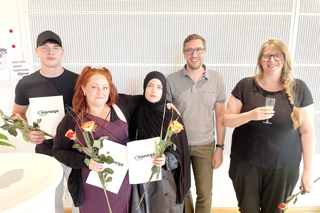 Fünf lächelnde Personen, drei mit "nanege"-Zertifikaten und Rosen, eine mit Getränk, feiern einen Erfolg.