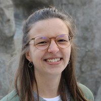 Nahaufnahme einer lächelnden jungen Frau mit Brille und langen braunen Haaren vor verschwommenem grauem Hintergrund.