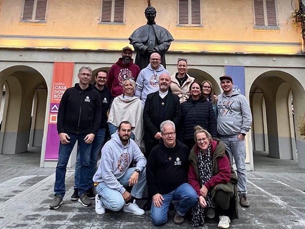 Eine Gruppe Menschen mit einem Priester posiert vor dem Don Bosco Museum mit einer Statue von Don Bosco darüber.