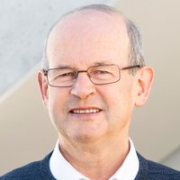 Kopfaufnahme eines älteren Mannes mit Brille, grauem Haar und blauem Pullover, der freundlich in die Kamera lächelt.