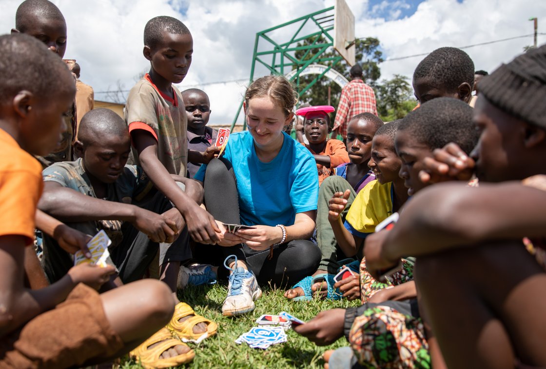 Eine junge Frau spielt mit einer Gruppe Kinder auf dem Gras Karten. Sie sind alle ins Spiel vertieft und lächeln.