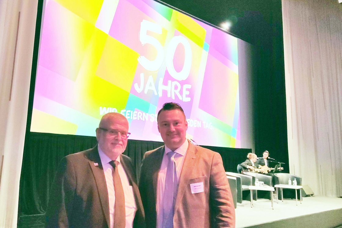 Zwei Männer in Anzügen lächeln vor einer großen Leinwand mit der Aufschrift „50 JAHRE“ auf einer Veranstaltung.