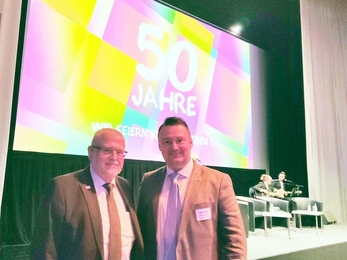 Zwei Männer in Anzügen lächeln vor einer großen Leinwand mit der Aufschrift „50 JAHRE“ auf einer Veranstaltung.