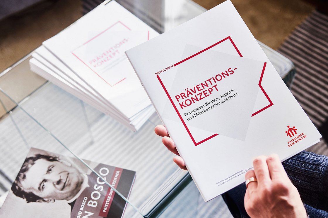 Eine Hand hält eine Broschüre mit dem Titel Präventionskonzept. Darunter liegen auf einem Glastisch weitere Broschüren mit Don Bosco. 