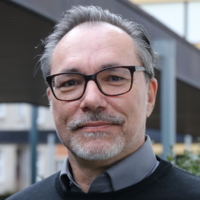 Lächelnder Mann mit grauen Haaren, Brille, Schnurrbart und Bart, im schwarzen Pullover über grauem Hemd im Freien.