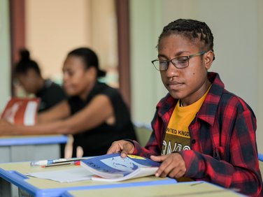 Eine Schülerin mit Brille und geflochtenen Haaren liest ein offenes Buch an einem Schultisch im Klassenzimmer.