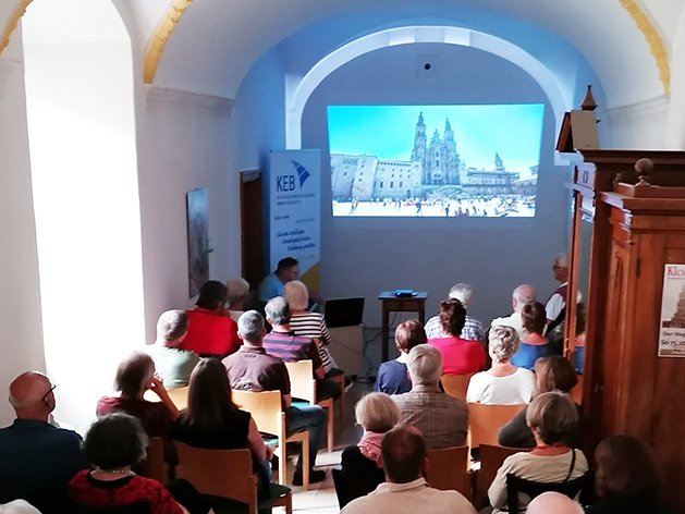 Publikum sieht Präsentation über den Jakobsweg, mit einem Bild der Kathedrale von Santiago de Compostela auf der Leinwand.