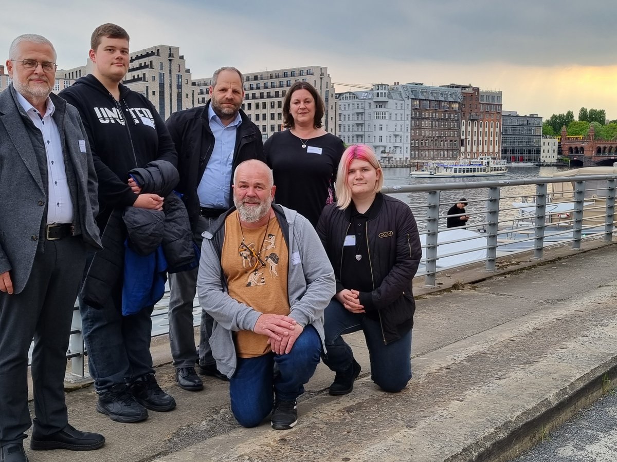 Sieben Menschen am Ufer eines Flusses, mit urbanen Gebäuden und der historischen Oberbaumbrücke im Hintergrund.