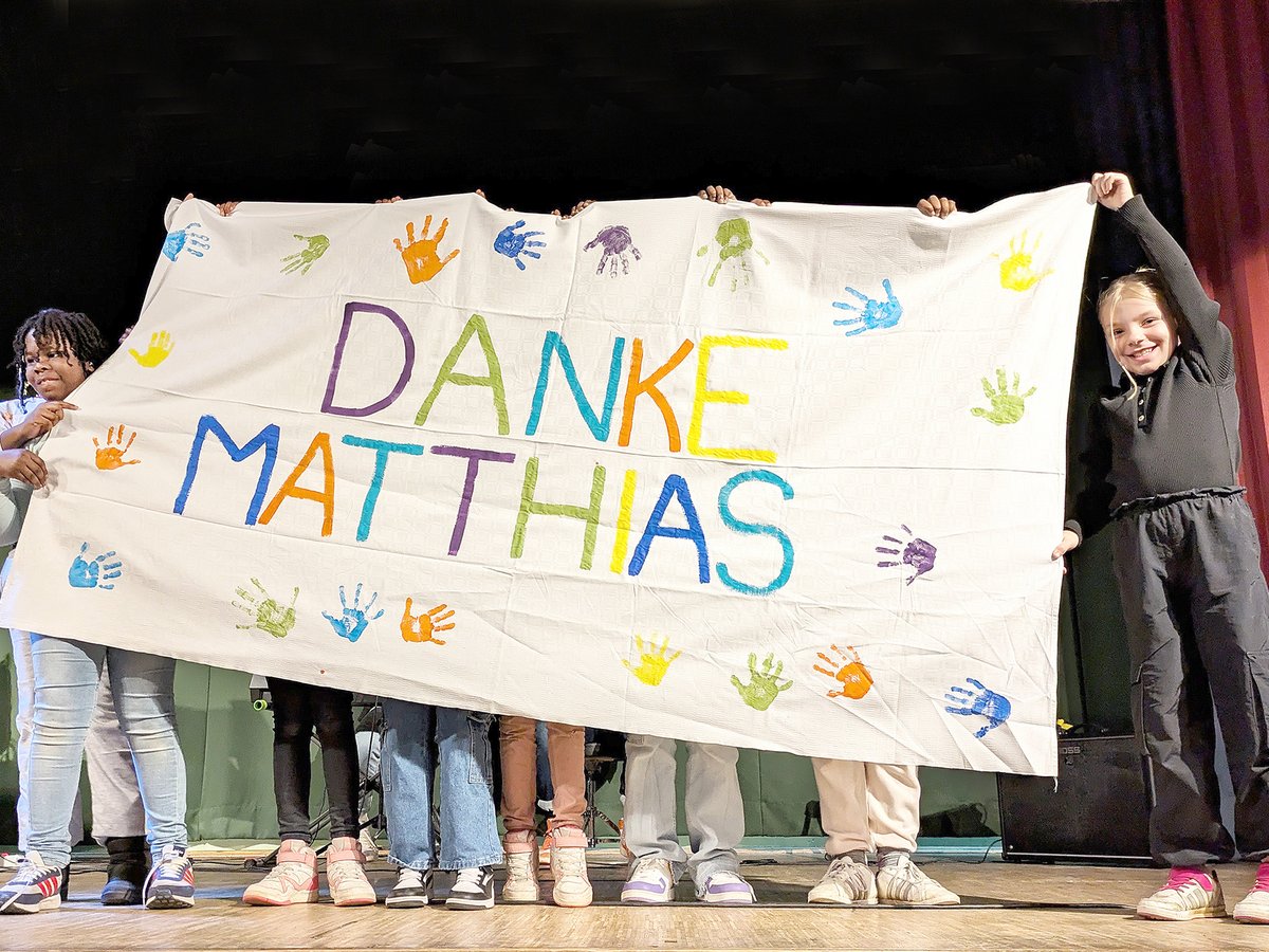 Kinder halten ein Banner mit der Aufschrift "DANKE MATTHIAS" und bunten Handabdrücken auf einer Bühne.