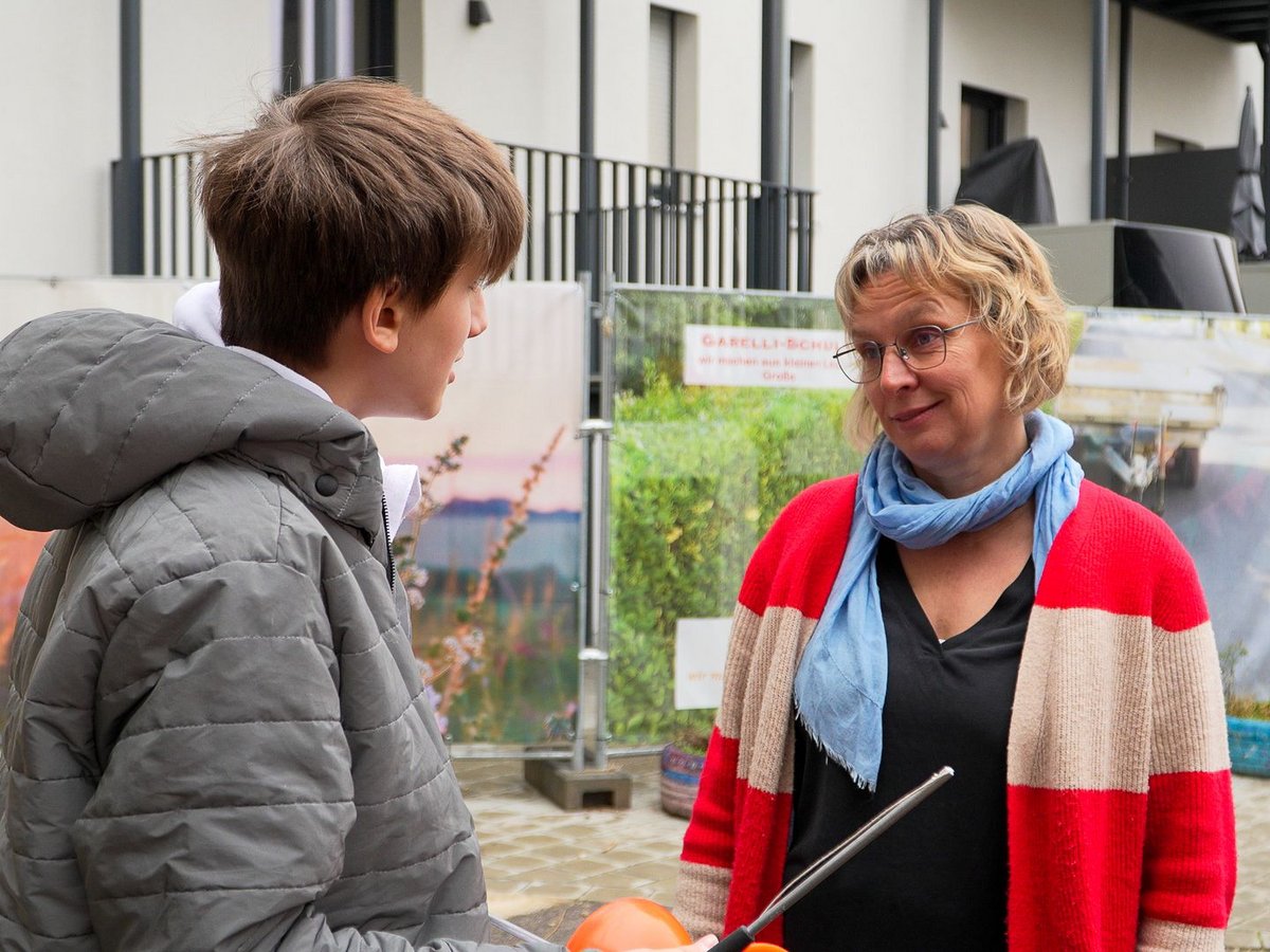 Ein junger Mensch in grauer Jacke spricht mit einer lächelnden Frau in rot-beiger Strickjacke und blauem Schal draußen.