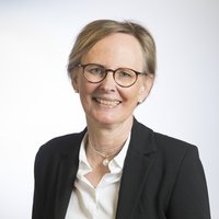 Lachende Frau mit Brille, hellbraunem Haar, schwarzem Blazer und Perlenkette vor weißem Hintergrund.