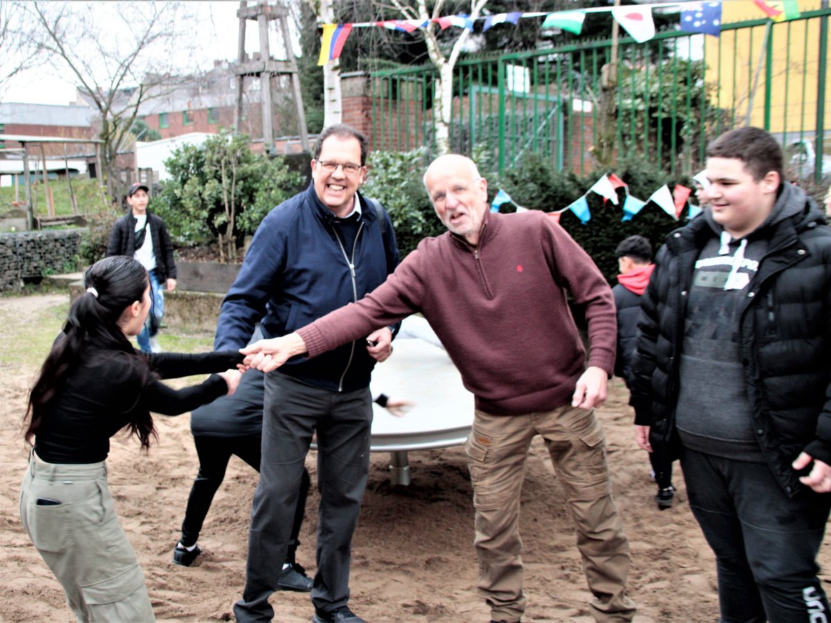 Eine lächelnde Gruppe von Menschen, die sich auf einem sandigen Spielplatz unter bunten Wimpeln die Hände schütteln.