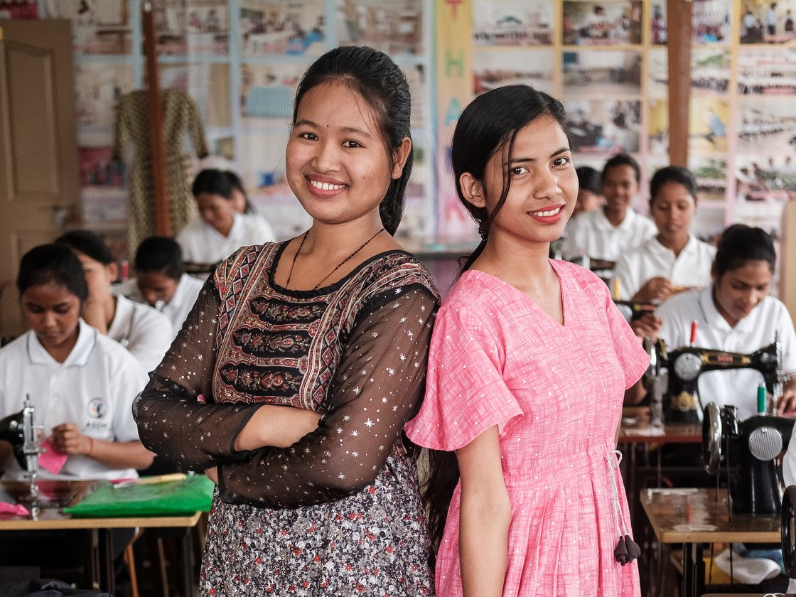 Zwei lächelnde junge Frauen stehen in einer Nähwerkstatt, mit anderen Schülern und Nähmaschinen im unscharfen Hintergrund.