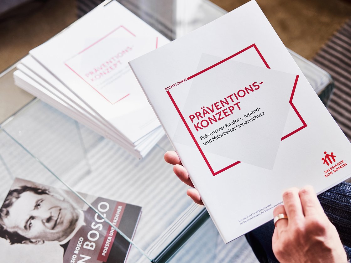 Eine Hand hält ein 'Präventionskonzept'-Heft für Kinder-, Jugend- und Mitarbeiterschutz. Stapel Hefte und ein Don Bosco Buch sichtbar.