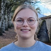 Lachende junge Frau mit blonden Haaren und runder Brille, draußen vor Bäumen und einem Gebäude.