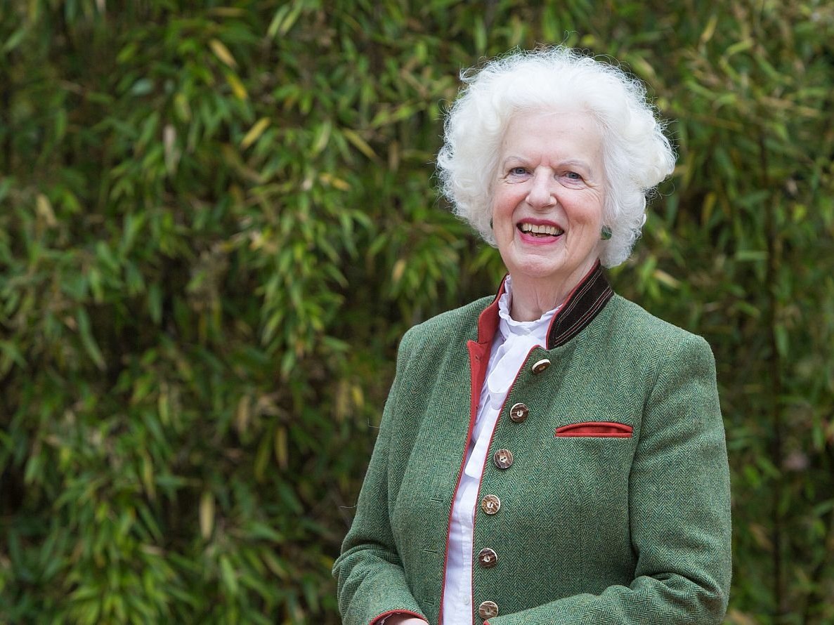 Lächelnde ältere Frau mit weißem Lockenhaar in grüner Jacke mit rotem Besatz, vor üppigem grünem Laub stehend.