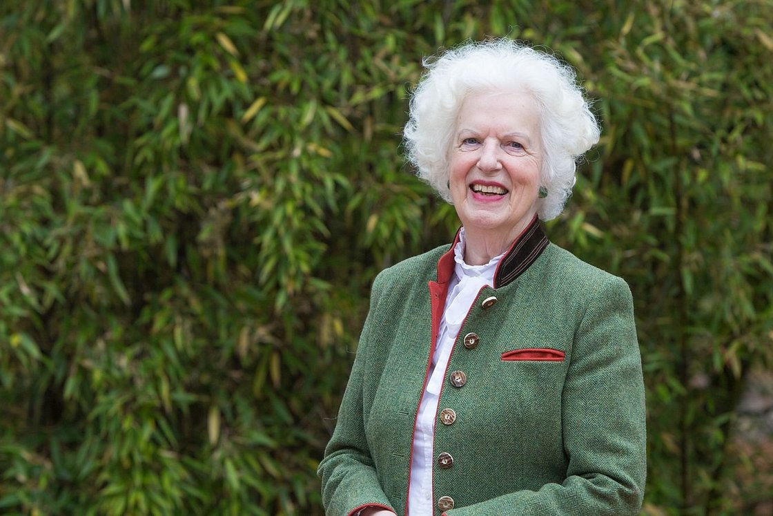 Lächelnde ältere Frau mit weißem Lockenhaar in grüner Jacke mit rotem Besatz, vor üppigem grünem Laub stehend.