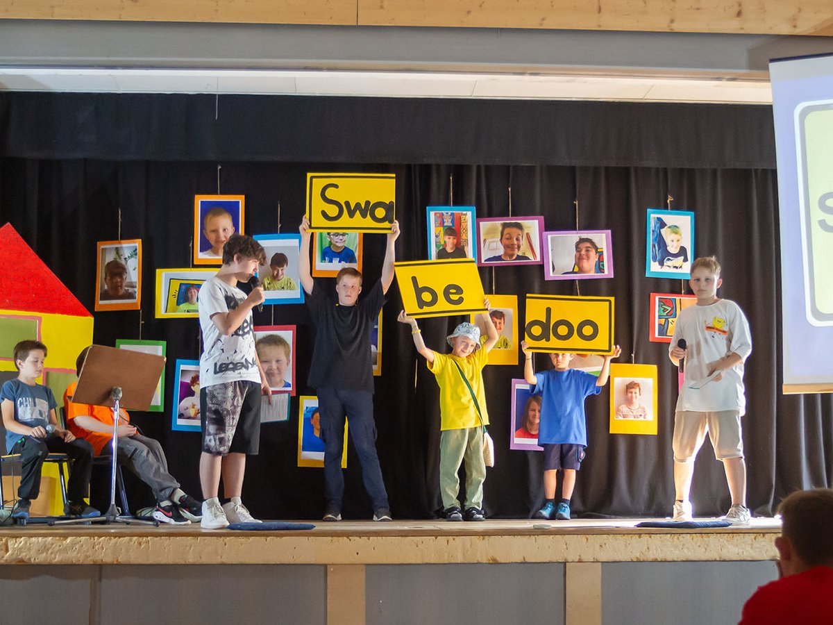 Kinder treten auf einer Bühne auf, einige halten gelbe Schilder mit 'Swa', 'be', 'doo'. Gerahmte Kinderfotos schmücken den Hintergrund.