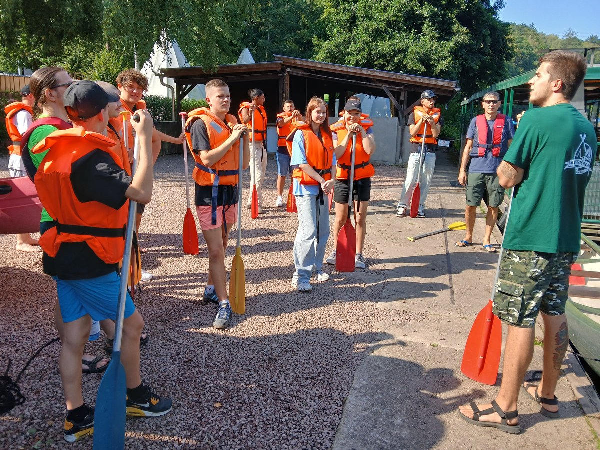 Eine Gruppe junger Menschen in orangefarbenen Schwimmwesten mit Paddeln, die einem Lehrer vor einer Kanutour zuhört.