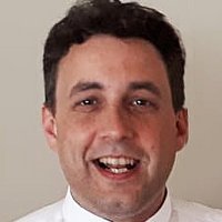 Ein lächelnder Mann mit kurzen dunklen Haaren, der ein weißes Hemd und einen Priesterkragen trägt.