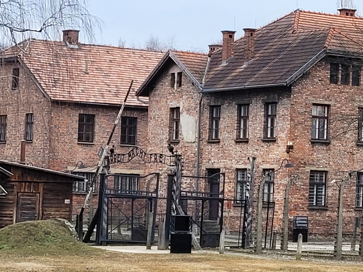 Das Eingangstor des Konzentrationslagers Auschwitz I mit dem Schriftzug „Arbeit Macht Frei“, dahinter Backsteingebäude und Stacheldraht.
