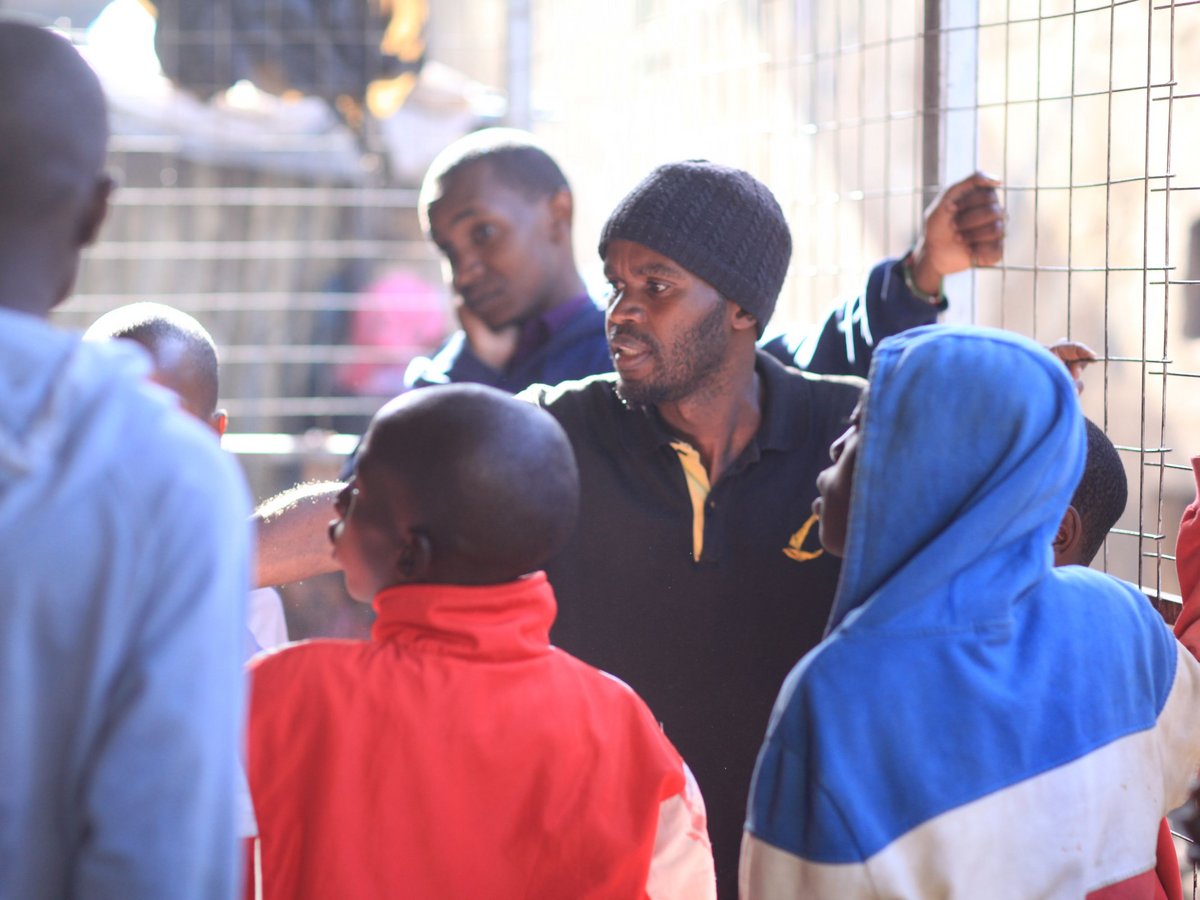 Eine Gruppe schwarzer Männer und Kinder versammelt, mit einem bärtigen Mann in schwarzer Mütze, der spricht. Kinder schauen ihn und ein Gitter an.
