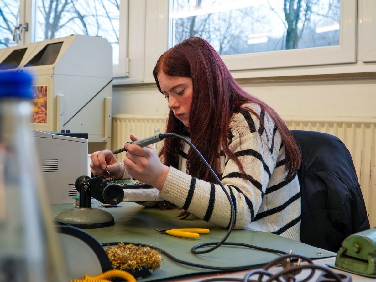 Eine konzentrierte junge Frau mit roten Haaren lötet eine Platine an einer Werkbank.