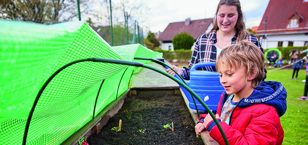 Junge und Mädchen pflanzen in einem Hochbeet mit grüner Netzabdeckung und betrachten neue Setzlinge.
