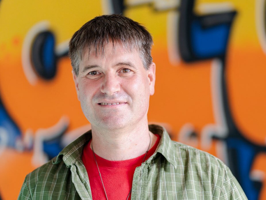 Ein lächelnder Mann mit kurzen braunen Haaren und einem grünen Karoshirt über einem roten T-Shirt vor einer unscharfen Graffiti-Wand.