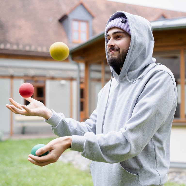 Ein lächelnder Mann in Kapuzenpullover und Mütze jongliert drei bunte Bälle (gelb, rot, grün) draußen im Garten.