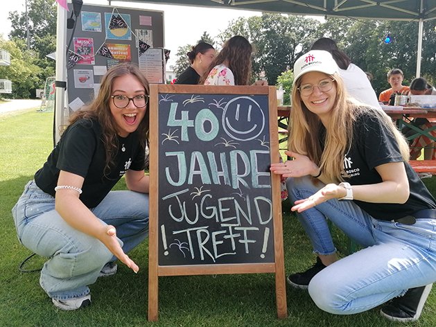 Zwei lächelnde junge Frauen zeigen auf eine Kreidetafel, die „40 Jahre Jugend Treff!“ bei einer Veranstaltung im Freien feiert.