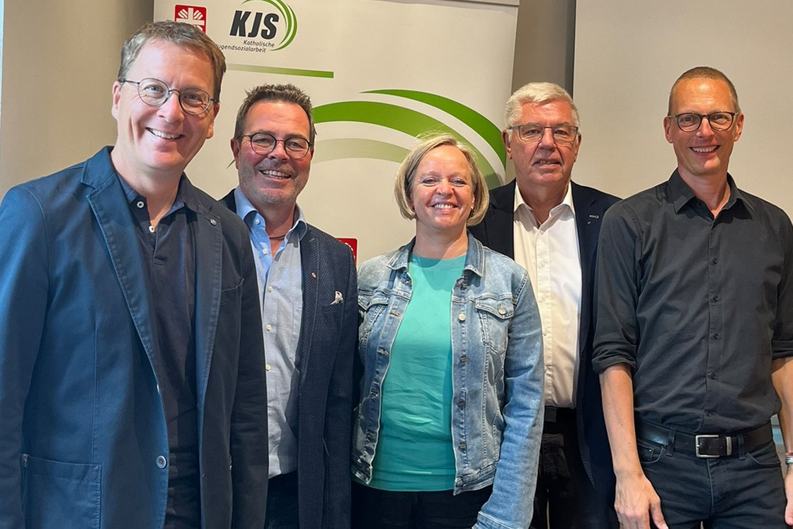 Fünf lächelnde Personen, vier Männer und eine Frau, vor einem Banner der Katholischen Jugendsozialarbeit (KJS).
