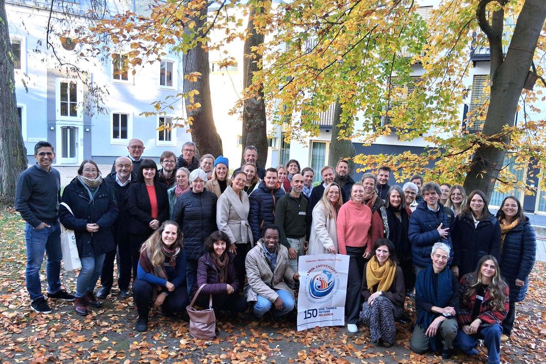 Vielfältige Gruppe lächelnder Menschen im Herbst im Freien, einige halten ein Schild 'Salesian Mission Day 2025'.