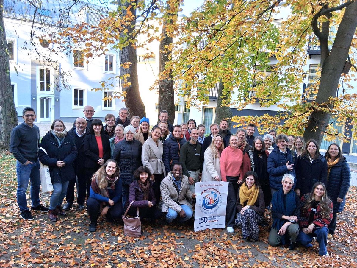 Vielfältige Gruppe lächelnder Menschen im Herbst im Freien, einige halten ein Schild 'Salesian Mission Day 2025'.