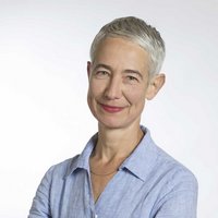 Porträt einer lächelnden Frau mit kurzen grauen Haaren, rotem Lippenstift und blauem Hemd vor weißem Hintergrund.